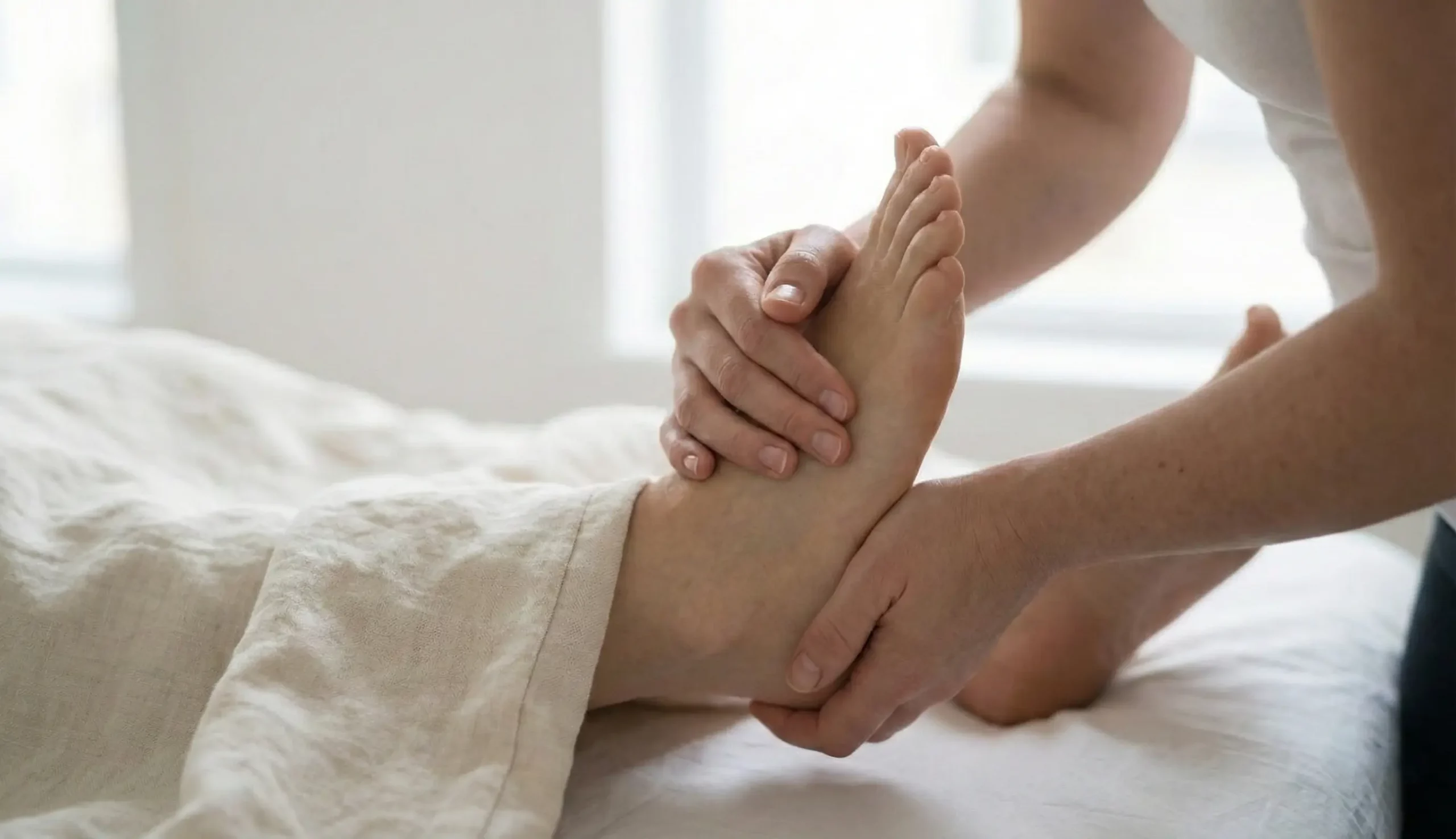 Pacjent leżący na plecach, przykryty kocem do kostek. Masażysta obiema rękami masuje prawą stopę.