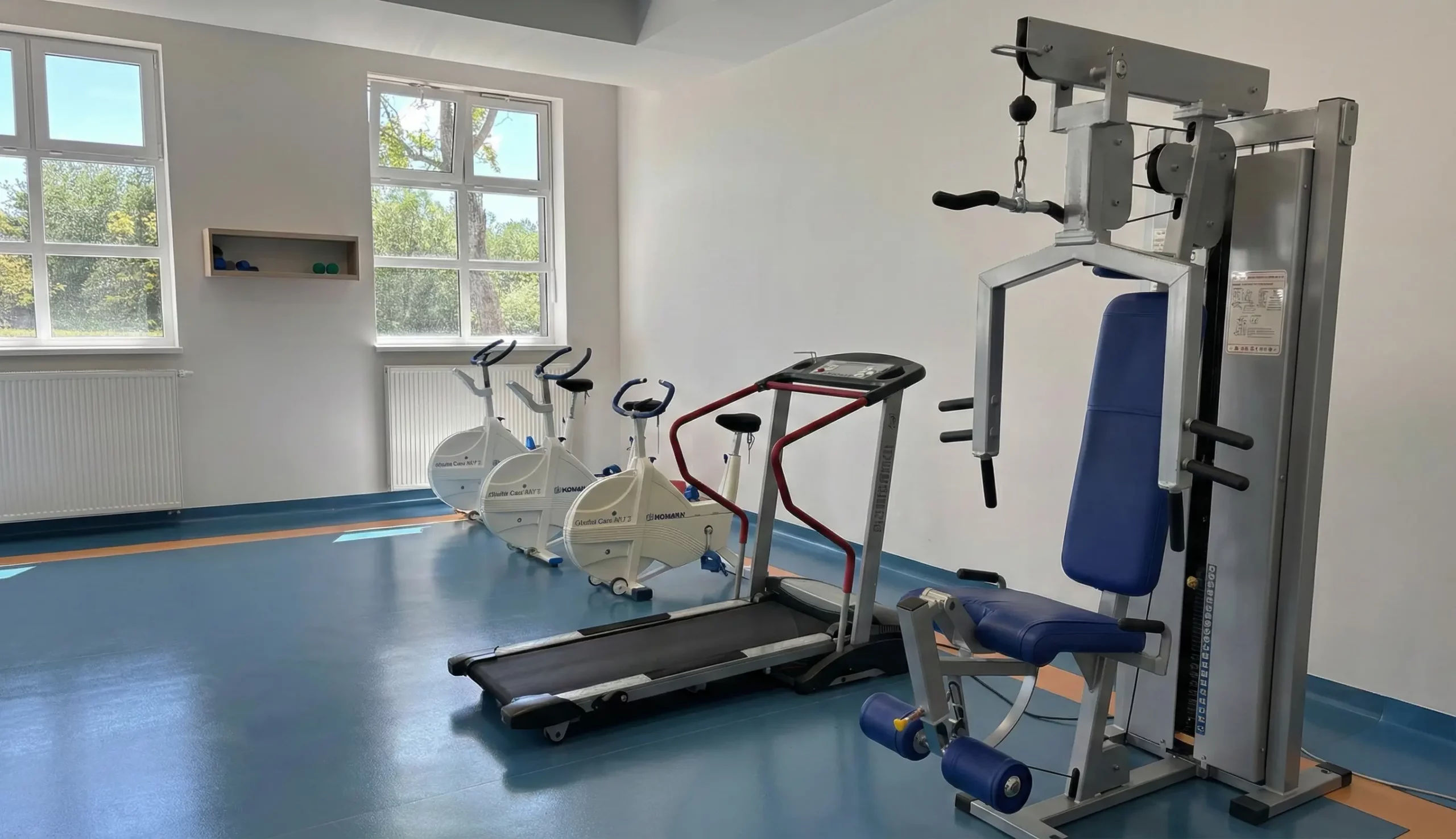 Sala gimnastyczna z widocznymi ergometrami, bieżnią i atlasem.